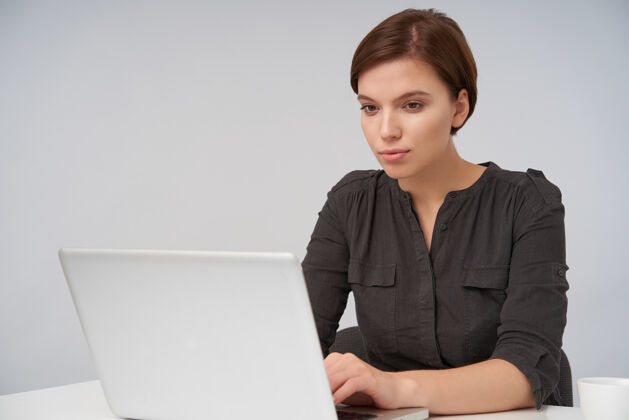 文字室内画像年轻的棕色头发可爱的女性工作与她的笔记本电脑 看着屏幕与平静的脸 穿着正式的衣服 而坐在白色摆姿势办公室正式