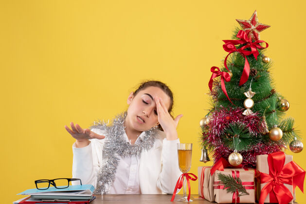 快乐正面图：女医生坐在黄色背景的桌子后面 还有圣诞树和礼品盒前面宝儿圣诞节