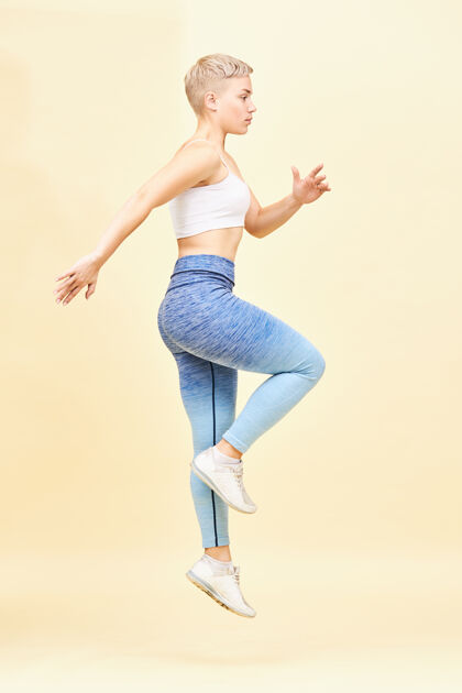 跳跃垂直侧视图全身充满活力的肌肉年轻的白人女运动员在顶部 紧身裤和运动鞋跳跃的地方 做有氧锻炼例行体型 减肥和活动体重女子健身房