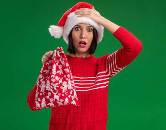 圣诞节遗憾的是年轻女孩戴着圣诞帽拿着圣诞礼物袋看着相机手放在头上隔离在绿色背景下头拿着袋子