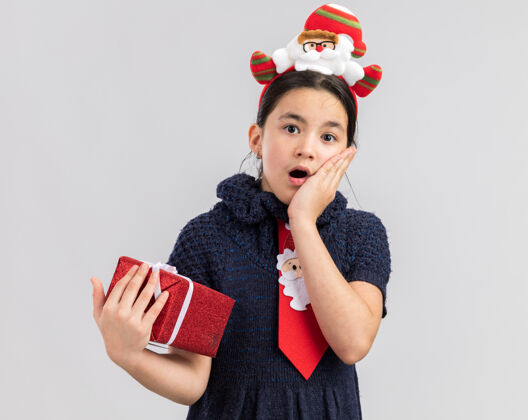 礼物小女孩穿着针织连衣裙 头上系着一条红色领带 头上戴着一个有趣的圣诞戒指 手里拿着圣诞礼物 看上去既惊讶又惊讶快乐小抱着
