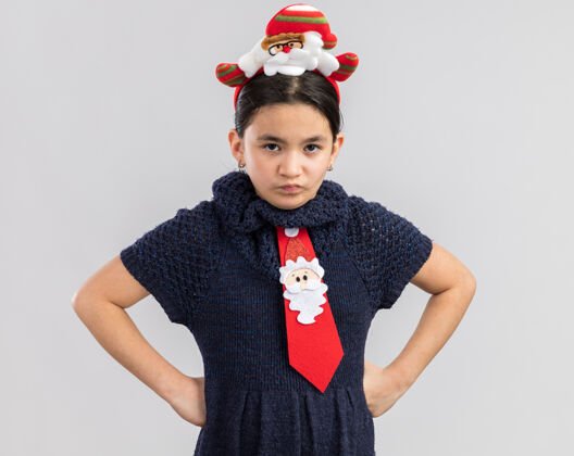 边缘小女孩穿着针织连衣裙 头上系着一条红色领带 头上戴着一个滑稽的圣诞边 眉头紧锁 胳膊放在臀部臀部新快乐