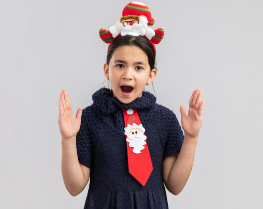 圣诞节小女孩穿着针织连衣裙 头上系着一条红色领带 戴着一个有趣的圣诞戒指 看上去很惊讶 用手做了一个大大的手势小手势搞笑