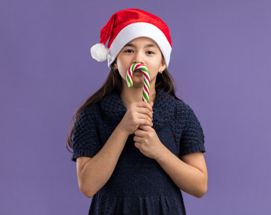 紫色小女孩穿着针织连衣裙 戴着圣诞帽 手里拿着糖果手杖 脸上洋溢着快乐和积极的笑容小站着圣诞