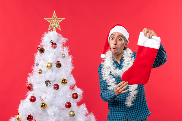 惊讶在红色圣诞树旁 一个戴着圣诞老人帽 穿着蓝色条纹衬衫 手拿圣诞袜子的年轻人大吃一惊帽子袜子年轻人
