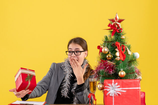 礼物前景色漫步的女孩戴着眼镜坐在桌子旁看着礼物 惊喜的圣诞树和鸡尾酒礼物流浪女孩圣诞节眼镜