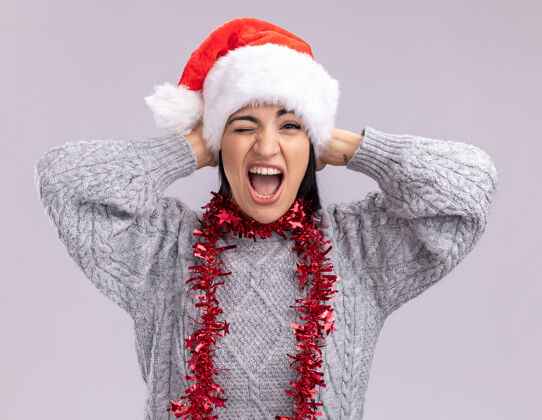 头快乐的年轻白人女孩戴着圣诞帽 脖子上戴着金箔花环 手放在头上 在白色的墙上尖叫和眨眼年轻周围脖子