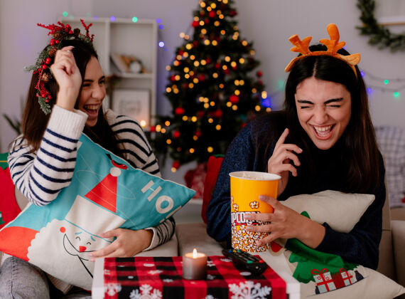 快乐带着冬青花环和驯鹿头带的快乐的年轻女孩们吃着爆米花 坐在扶手椅上玩耍 在家里享受圣诞节时光年轻爆米花圣诞快乐