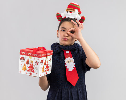新小女孩穿着针织连衣裙 头上系着一条红色领带 头上戴着一个有趣的圣诞圈 手里拿着圣诞礼物 看起来很开心 很积极 通过ok标志看抱着搞笑衣服