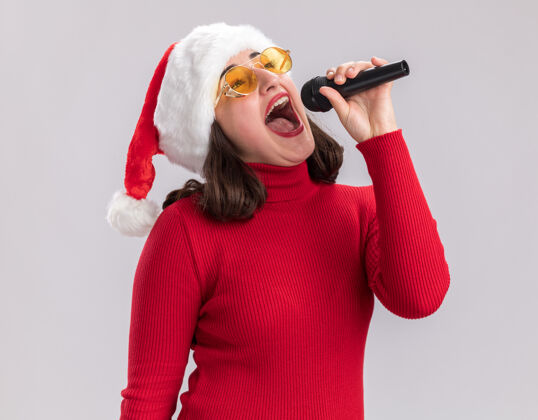 毛衣快乐快乐的年轻女孩穿着红毛衣 戴着圣诞帽 戴着眼镜 手持麦克风 站在白墙上唱着一首歌唱歌站着唱歌