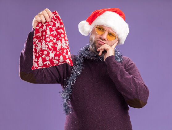 圣诞节困惑的中年男子戴着圣诞帽 脖子上戴着金箔花环 戴着眼镜 手放在下巴上 看着圣诞礼物袋 手被隔离在紫色的墙上中年脖子保管