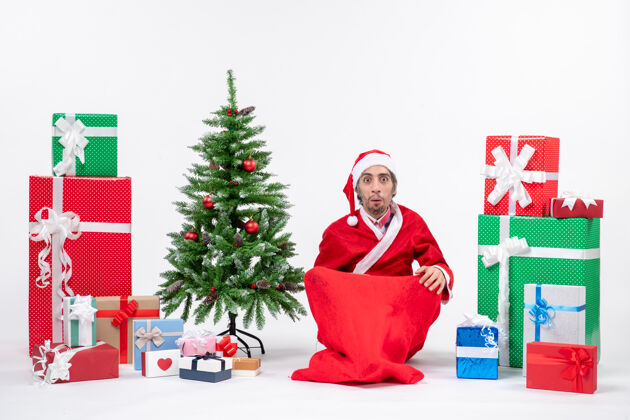 年轻人年轻人打扮成圣诞老人 拿着礼物和装饰好的圣诞树坐在地上寻找什么装饰成人圣诞老人