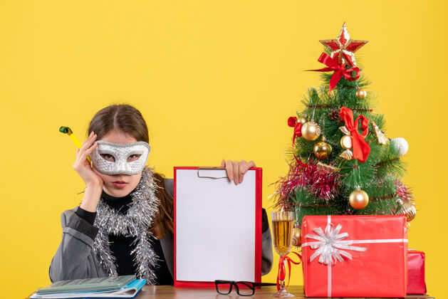 严肃前视图严肃的年轻女孩戴着面具坐在桌子旁拿着噪音制造者圣诞树和鸡尾酒礼物丝带礼物桌子