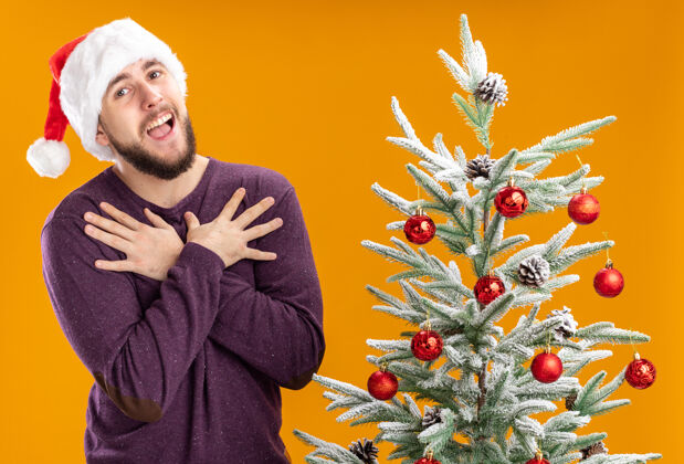 相机身穿紫色毛衣 戴着圣诞帽的年轻人看着相机 脸上挂着幸福的手 胸前放着橙色背景下的圣诞树年轻人手男人
