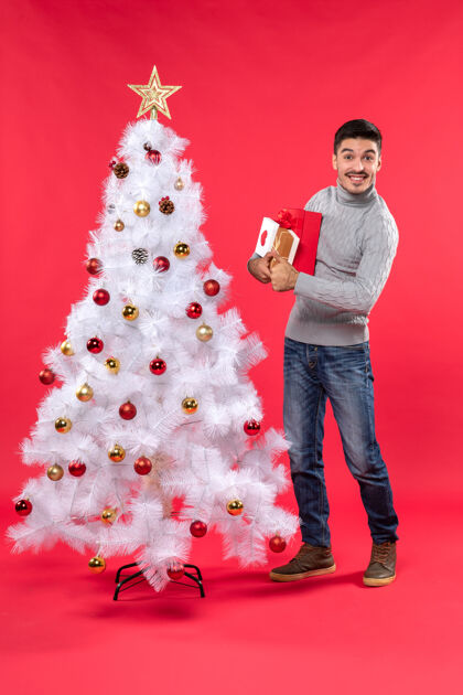 年轻人英俊的年轻人微笑着站在装饰过的白色新年树旁 手里拿着他的礼物帅哥庆祝圣诞