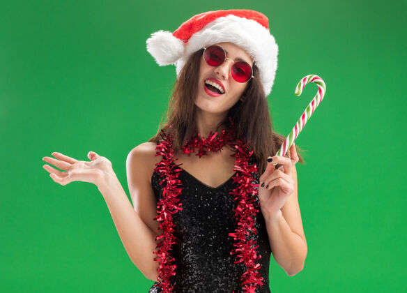 快乐快乐的年轻漂亮女孩戴着圣诞帽戴着眼镜脖子上戴着花环拿着圣诞糖果摊开手孤立在绿色的墙上花环年轻帽子