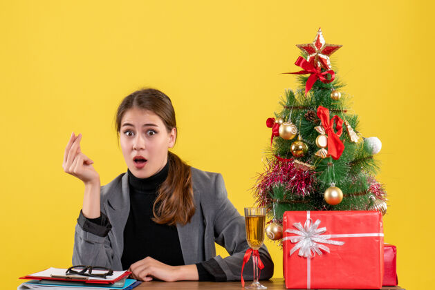 惊讶的女孩正面图惊讶的女孩坐在桌子前展示金钱手势圣诞树和鸡尾酒礼物手势鸡尾酒冬天