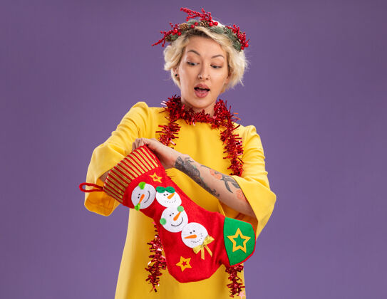 金发好奇的金发女郎头戴圣诞花环 脖子上戴着金箔花环 手里拿着圣诞长袜 把手伸进袜子里 看着孤立在紫色墙上的复制品放年轻金属丝