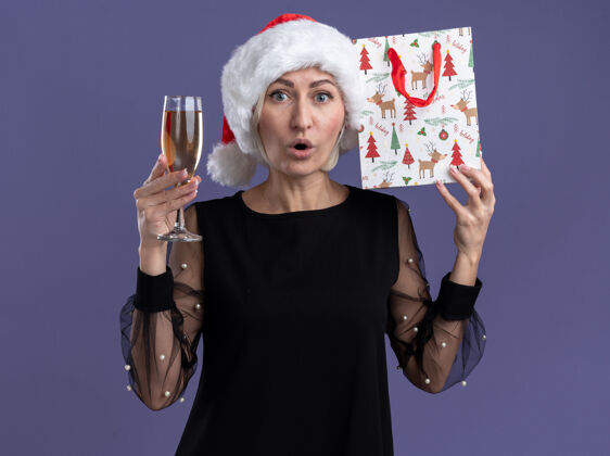 头惊讶的中年金发女人戴着圣诞帽看着相机拿着一杯香槟和触摸头部与紫色背景上孤立的圣诞礼品袋礼物香槟惊讶