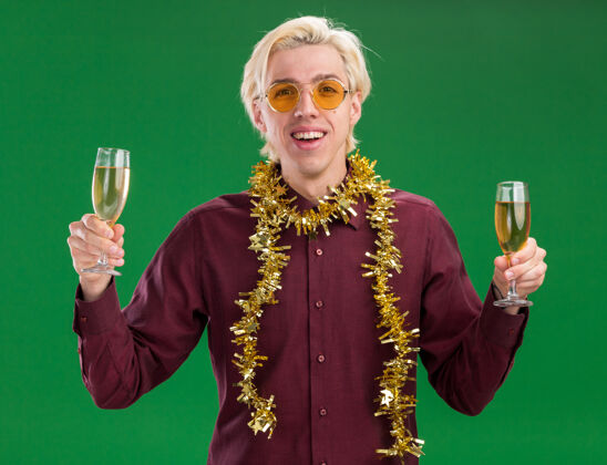 金箔快乐的年轻金发男子戴着眼镜 脖子上戴着金箔花环 手里拿着两杯香槟 隔离在绿色的墙上花环金发穿着