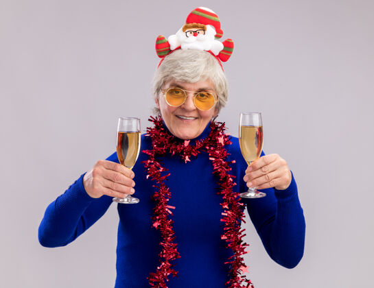 新年戴着太阳眼镜 脖子上戴着圣诞老人头带和花环 面带微笑的老妇人 手里拿着一杯香槟酒 背景是白色的 还有复印空间圣诞老人眼镜老人