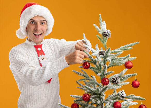 站着令人印象深刻的年轻帅哥戴着圣诞帽和圣诞老人领带站在圣诞树附近装饰圣诞球装饰隔离在橙色的墙上近年轻球