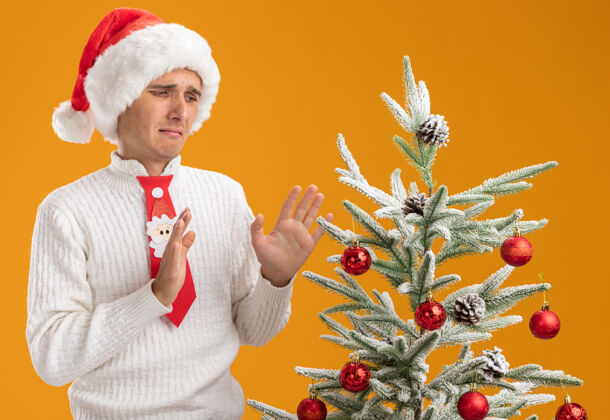 圣诞老人年轻帅哥戴着圣诞帽 打着圣诞老人的领带 站在装饰好的圣诞树旁 看着它做着拒绝的手势 孤立在橙色的背景上看帽子手势