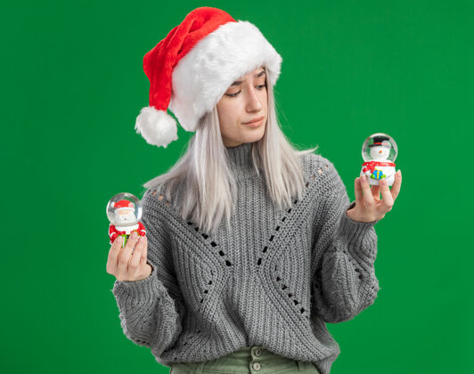 举行年轻的金发女郎穿着冬季毛衣 戴着圣诞帽 手里拿着圣诞玩具雪球 站在绿色的背景下 困惑地试图做出选择使站着圣诞老人