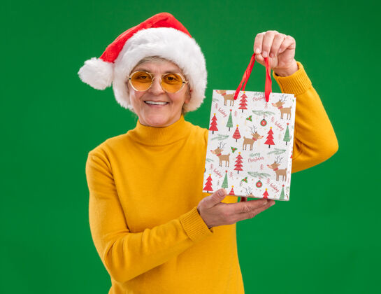 拿着微笑的老太婆戴着太阳眼镜 戴着圣诞老人的帽子 手里拿着一个纸礼包 背景是绿色的 有复印空间微笑眼镜圣诞老人