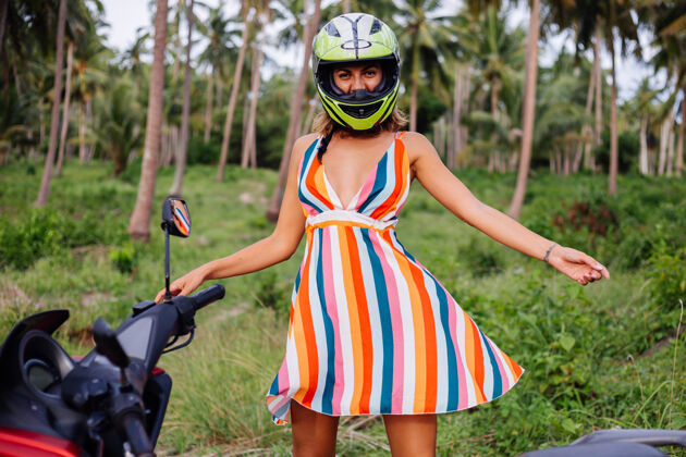 光明快乐的高加索骑摩托车的女人 穿着五颜六色的夏装 戴着摩托车头盔度假骑热带旅游