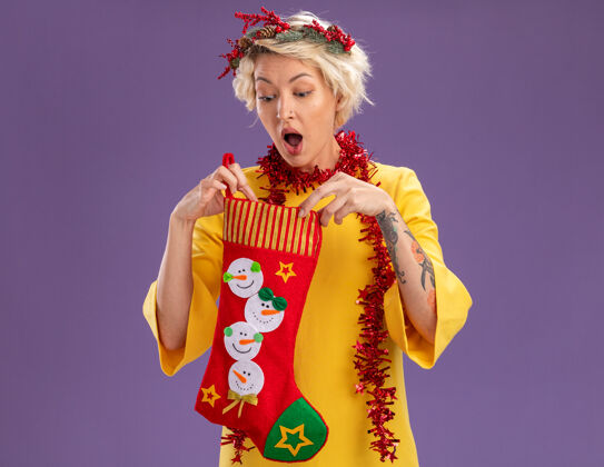 女人好奇的金发女郎头戴圣诞花环 脖子上戴着金箔花环 手里拿着圣诞长袜 看着里面 紫色的墙上有一个单独的复制空间圣诞节花环年轻