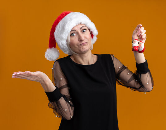 金发笨手笨脚的中年金发女人戴着圣诞帽拿着圣诞老人的圣诞饰品 空手孤立在橙色的墙上女人圣诞老人无知