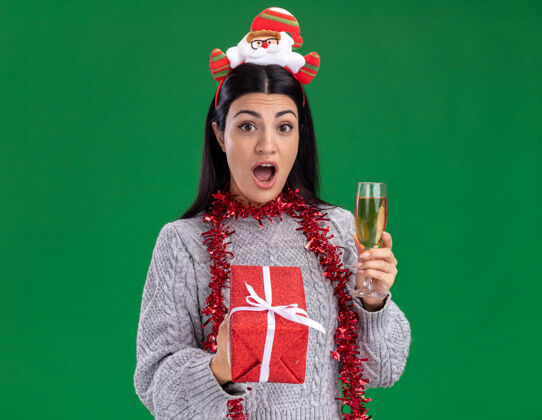 周围令人印象深刻的年轻白人女孩戴着圣诞老人的头带和金箔花环在脖子上举行的礼品包和一杯香槟隔离在绿色墙上的复制空间玻璃圣诞老人头带
