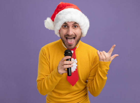 帽子顽皮的白人年轻人戴着圣诞帽 打着领带 拿着麦克风 看着摄像机 镜头上的舌头在做摇滚 背景是紫色的 有复制空间看年轻圣诞快乐