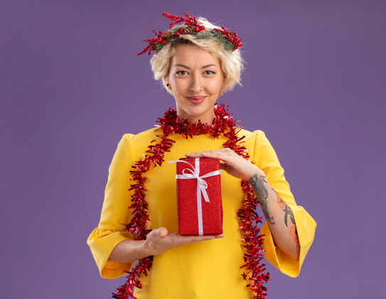 头部年轻的金发女郎戴着圣诞花环 脖子上戴着金箔花环 手里拿着礼包 看着紫色背景上的相机包装圣诞节花环