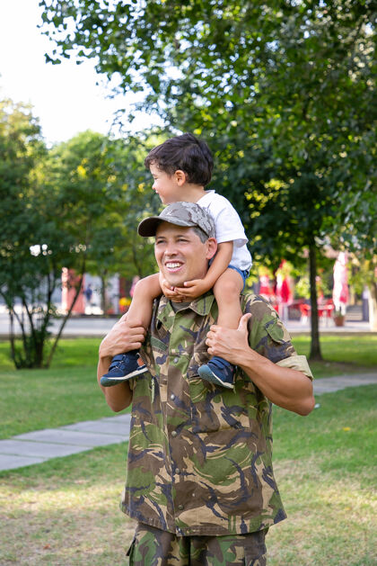 回归快乐的爸爸抱着儿子的脖子 抱着他的腿快乐的白人爸爸穿着制服和小男孩在公园散步可爱的男孩看着别处家庭 父亲和回家的概念父母爸爸士兵