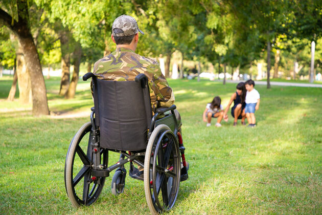 男人孤残退役军人坐在轮椅上看着妻子和小孩在公园里一起玩耍后视图老兵的战争或残疾概念女儿士兵孩子