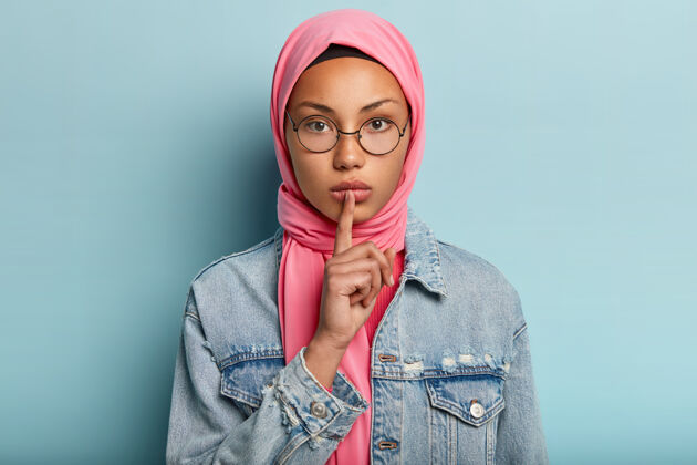 沉默一位穿着粉色围巾 牛仔外套的严肃女性头像 用食指捂住嘴唇 要求不要吵闹 做出安静的手势 有伊斯兰信仰 戴圆眼镜安静严肃手势