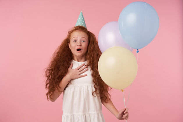 女性快乐的木发红发小女孩手持气球站在粉色背景上 被客人逗乐 开心地笑着孩子和庆祝的概念室内女孩礼物