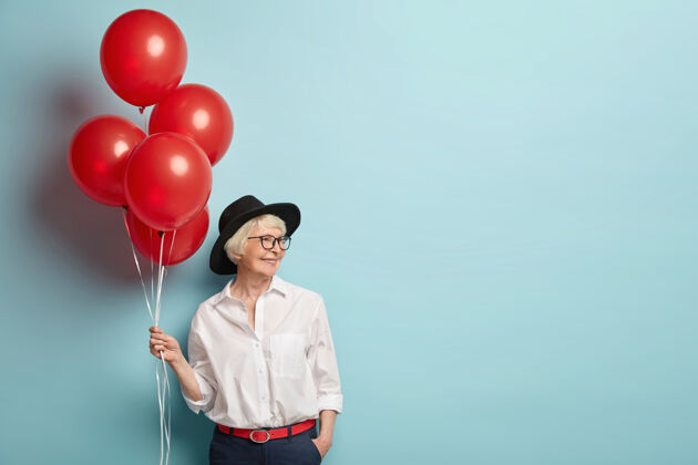 老年人喜笑颜开的女士 皮肤有皱纹 在工作中和同事聚会 庆祝退休 手持红色气球 穿着时髦的衣服 隔着空白的蓝色墙壁与世隔绝高兴时尚快乐