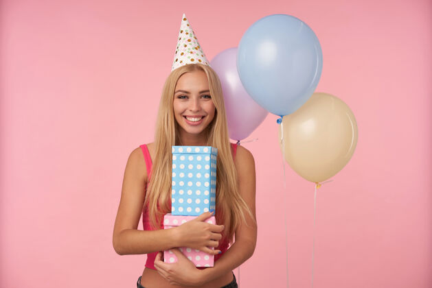帽子迷人的正面金发女性的肖像 长发手持礼品包装盒 展示她愉快的情绪 在节日服装的粉色背景下摆姿势盒子空气头发