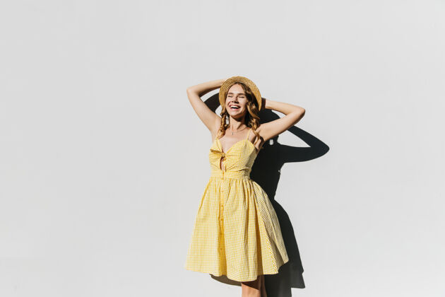 阳光美丽苗条的戴帽子的女士在阳光明媚的日子里摆姿势大笑穿着黄色连衣裙的幽默女模特的户外镜头女性高兴模特