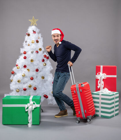 灰色年轻人兴高采烈地穿着灰色的红色旅行箱走着购物圣诞树帽子
