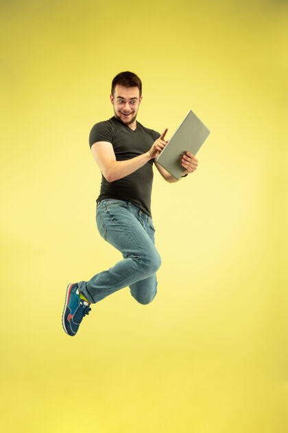 连接快乐跳跃的人与黄色背景上隔离的小玩意全长肖像现代技术 自由选择的概念 情感的概念使用笔记本电脑的工作和飞行乐趣男孩办公室运动