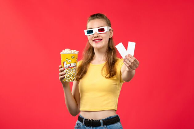 视图正面图年轻女性戴着d墨镜在电影院里拿着电影票和爆米花在红墙电影院里女性的颜色票电影院成人