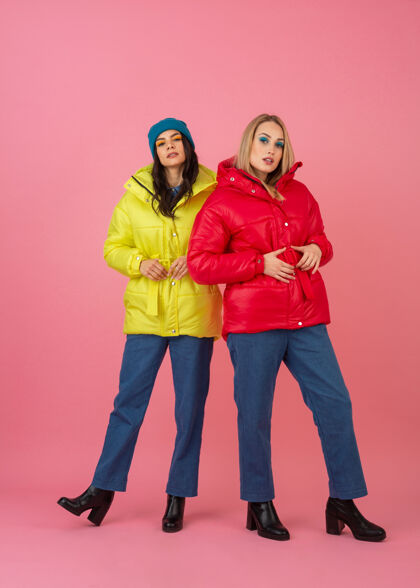 衣服两个迷人的女孩在粉红色的背景下 穿着鲜艳的红色和黄色的彩色冬季羽绒服摆姿势时尚粉色户外