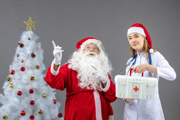 第一圣诞老人和女医生的前视图 女医生拿着急救箱在灰色的墙上前面援助圣诞老人