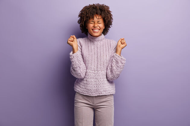 牙齿乐观的非裔美国女人的孤立镜头 快乐地握紧拳头 积极地微笑 快乐地闭上眼睛 穿着时髦的服装 站在紫色的墙上成功和胜利表情卷发胜利