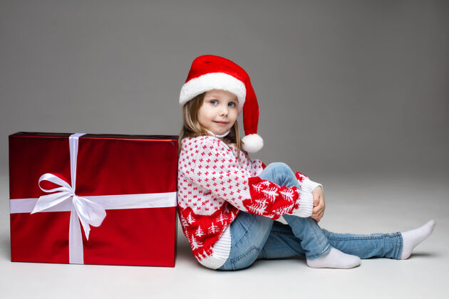 毛衣小女孩戴着圣诞帽 穿着冬季图案的毛衣 斜靠在白色蝴蝶结的红色圣诞礼物上灰色墙上的摄影棚拍摄可爱风景室内