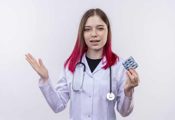 白色高兴的年轻医生女孩穿着听诊器医用长袍拿着药片在孤立的白色背景上摊开手穿着传播听诊器